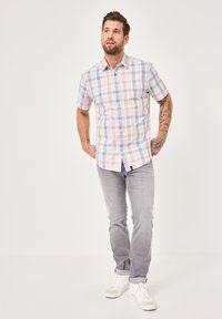 Kurzärmliges kariertes Hemd in Pastellfarben mit Knopfleiste, kombiniert mit hellgrauen Jeans und weißen Sneakers. Tätowierungen sind am Arm sichtbar.