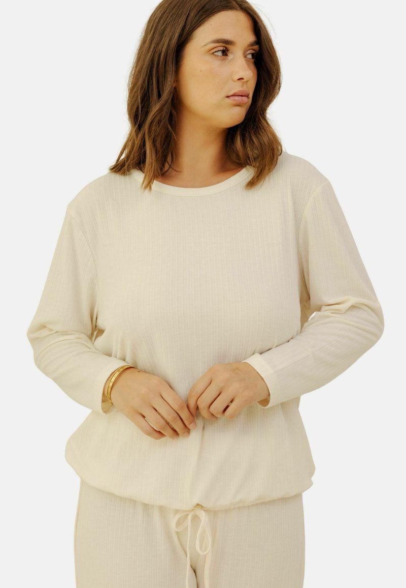 Haut crème à manches longues avec une texture côtelée et un col rond. Présente un ourlet élastique et une coupe décontractée. Associé à un pantalon à cordon assorti.