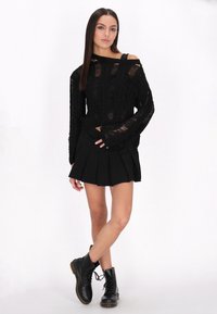 Schwarzer gestrickter Pullover mit offenen Schultern, der ein strukturiertes Muster aufweist. Kombiniert mit einem schwarzen plissierten Rock und schwarzen Schnürkampfstiefeln.