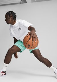 Atlet, der bærer en hvid t-shirt med sort logo, mintgrønne shorts med hvide detaljer og sorte og hvide sneakers, dribler en basketball.