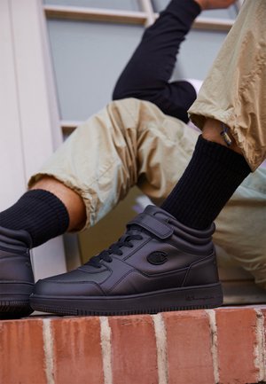 Sneakers neri alto in pelle, con una fascia in velcro, dettagli testurizzati e design con lacci. Indossati con calze nere a coste e pantaloni beige.