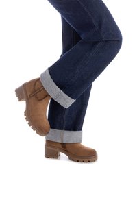 Stivali alla caviglia in suede marrone con tacco largo, abbinati a jeans di denim scuro arrotolati con risvolti grigio chiaro. Superficie testurizzata sugli stivali.