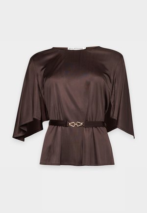 Une femme porte un chemisier marron ceinturé, une jupe midi noire et des sandales noires à talons, sur un fond clair uni.