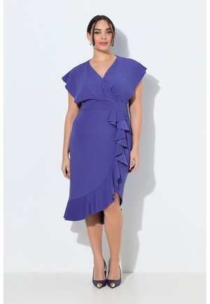 Kvinde iført en knælængde lilla slå-om kjole med flæsedetaljer og kappylder, kombineret med mørke spidse hæle, stående foran en hvid væg.