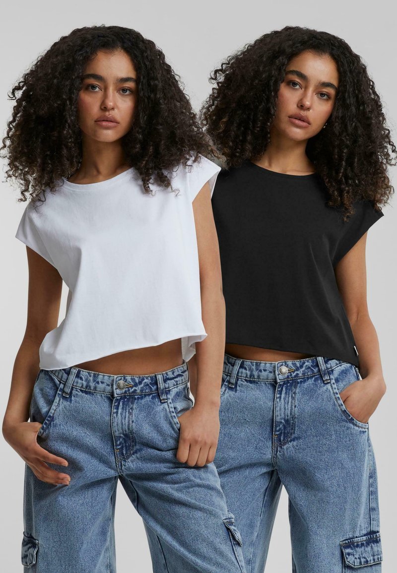 Félvállas fehér és fekete pamut cropped pólók, rövid ujjakkal. Magas derekú, világoskék farmernadrággal párosítva, amely különböző zsebekkel rendelkezik.