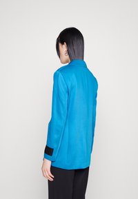 Turkusowa marynarka z klasycznym kołnierzem, usztywnianymi ramionami i czarnymi mankietami. Gładka faktura i dopasowany krój. Jednolite tło.