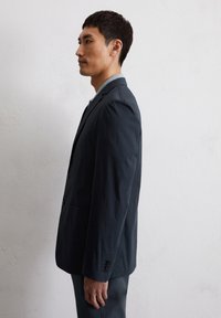Granatowa marynarka o fakturowanej tkaninie, zapinana na jeden guzik, z dwoma przednimi kieszeniami i mankietami na guziki. Model zaprezentowany w profilu.