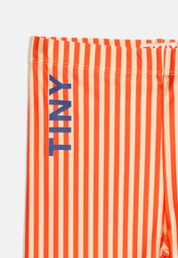 Shorts rayés verticaux orange et beige avec le mot "TINNY" imprimé en bleu sur le côté gauche.