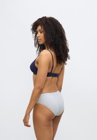 Σκούρο μπλε σουτιέν με λεπτές τιράντες, υφασμένο με υφή. Ανοιχτό μπλε ψηλόμεσο εσώρουχο με διακοσμητική δαντέλα. Η μοντέλο ποζάρει πλάγια μπροστά σε λευκό φόντο.