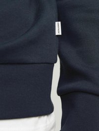 Sudadera azul marino con puños y bajo acanalados, que presenta una pequeña etiqueta blanca con "JACK & JONES". Tejido de algodón suave, de textura lisa.
