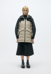 Καφέ και μαύρο φουσκωτό μπουφάν με κουκούλα, χαρακτηρισμένο από επενδυμένες ραφές, δύο μπροστινές τσέπες και πλήρη φερμουάρ. Φοριέται πάνω από μια μαύρη φ�ούστα και παπούτσια.