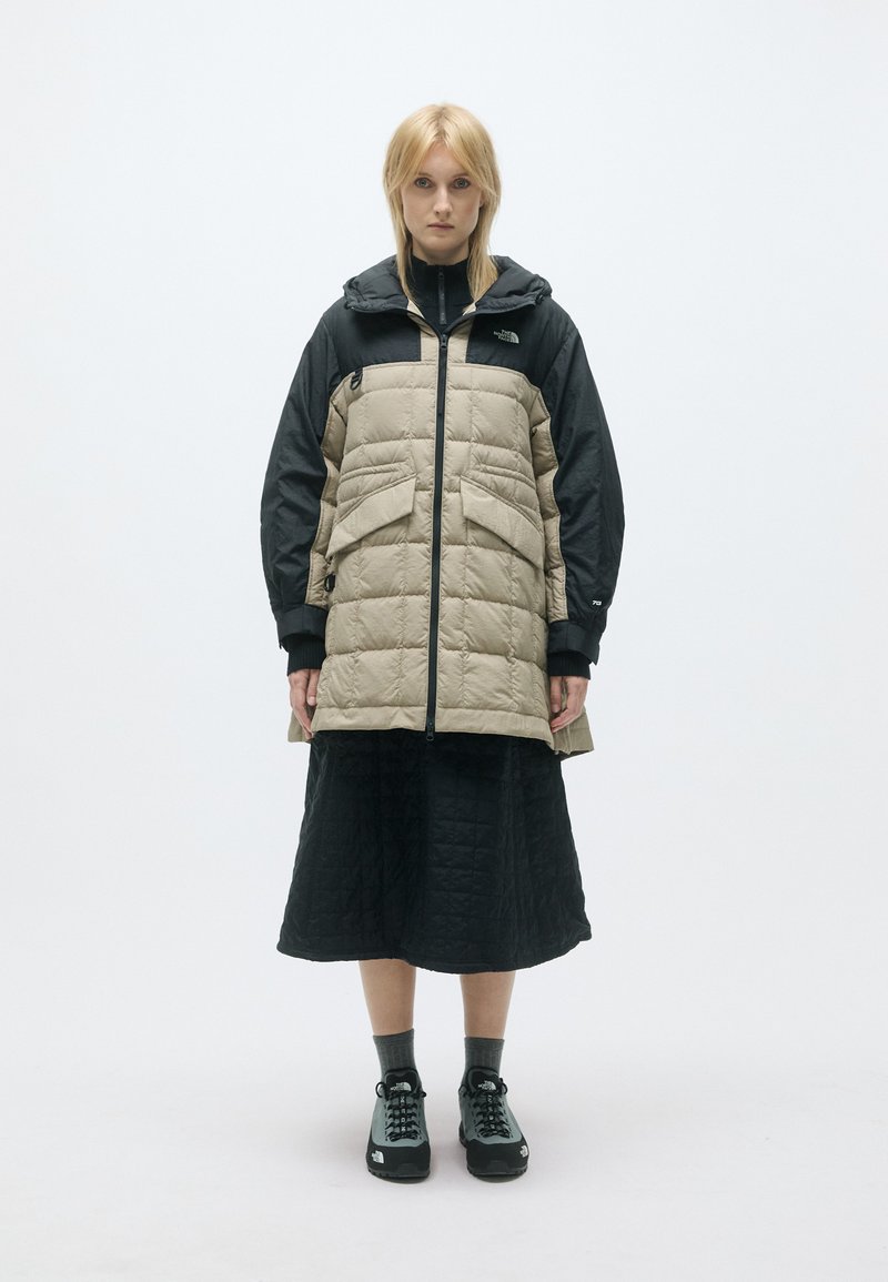 Chaqueta acolchada en tonos marrón y negro con capucha, presenta un relleno acolchado, dos bolsillos frontales y una cremallera completa. Lucida sobre una falda negra y calzado.