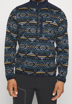 Marineblauwe pullover met geometrisch patroon in turquoise, geel en wit. Kenmerkt zich door een kraag met halve rits en effen marineblauwe schouderdetails.