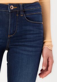 Dark blue jeans made of denim with orange stitching, featuring a standard five-pocket design, and a copper button at the waist.