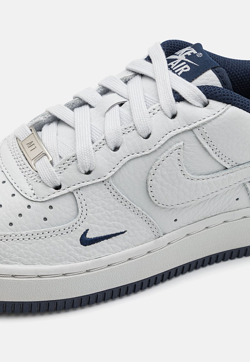 Zapatilla blanca de cuero con acentos en azul marino, puntera perforada y cordones blancos. Presenta un acabado texturizado y un logotipo de la marca visible en la lengüeta.