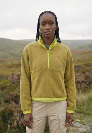 Jeune femme aux cheveux tressés portant une veste polaire verte et un pantalon beige, debout en plein air dans un paysage vallonné et herbeux.