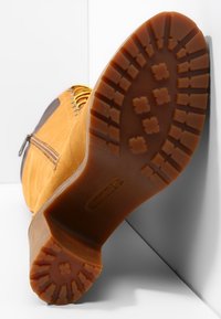 Bota de cuero marrón con una suela de goma texturizada, que presenta un patrón de zigzag y cordones amarillos. Cremallera lateral para facilitar el uso.
