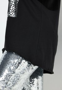 Zwarte t-shirt met een metallic slangenhuidpatroon op de mouw, gecombineerd met glanzende zilveren paillettenbroek, wat een gestructureerd, reflecterend oppervlak laat zien.