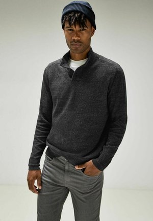 Dunkelgrauer Strickpullover mit einem Viertel-Reißverschluss-Kragen und gerippten Bündchen. Kombiniert mit hellgrauen Slim-Fit-Hosen trägt das Modell eine marineblaue Mütze.