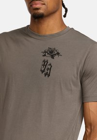 T-shirt grigio a maniche corte con una grafica di rosa nera e testo stilizzato "U/A" sul petto. Materiale morbido in cotone con scollatura a girocollo.