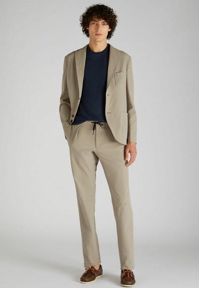 Costume beige texturé avec une veste et un pantalon à cordon, associé à un T-shirt bleu foncé et des chaussures marron. Design ajusté, accents minimalistes.