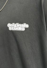 Mørkegrå bomulds T-shirt med et hvidt "Originals Studio" logo på venstre brystområde, lavet af et glat, struktureret stof.