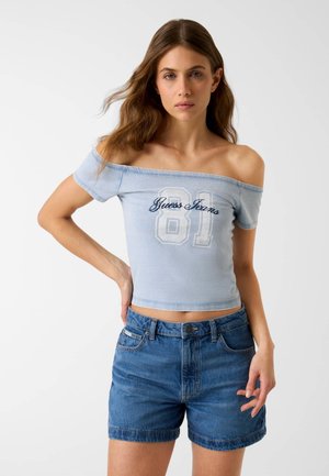 Jeune femme aux longs cheveux bruns portant un haut bleu clair à épaules dénudées avec l'inscription "Guess Jeans 81" et un short en denim, debout devant un fond uni.