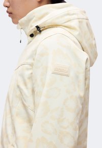 Personne portant une veste beige clair à motif avec une capuche et un patch logo Icepeak sur la partie supérieure du bras.