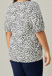 Mujer vestida con una blusa de manga corta blanca y negra con flores, combinada con pantalones azul oscuro, vista desde atrás contra un fondo sencillo.