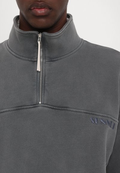 Sudadera de forro polar gris con cuello alto, media cremallera y logo bordado en azul marino. Textura suave con costuras y puntadas visibles.