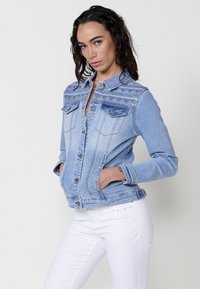 Világoskék farmerdzseki mintás részletekkel, gombos záródással, zsebekkel a mellrészen és karcsú fazonnal. Hosszú ujjú, diszkrét textúrával rendelkezik.