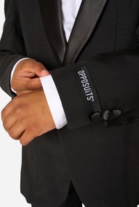 Must ülikonnajakk satiinklappel, valge triiksärgi mansetid paistmas, millel on nööbiaksendid ja OPPOSUITS silt varrukal.