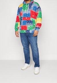 Wielokolorowa bluza z kapturem z wzorem patchworkowym, w którym znajdują się wzory bandany w kolorach niebieskim, czerwonym, zielonym, żółtym i białym, zestawiona z niebieskimi dżinsami i białymi sneakersami.