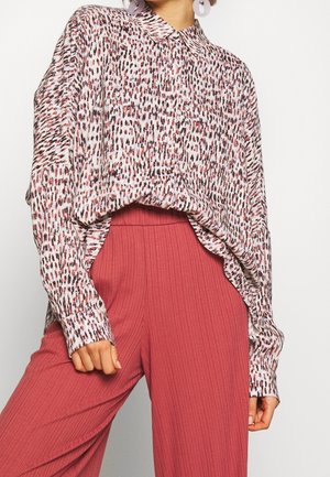 Femme portant un chemisier à manches longues à motif, rentré dans un pantalon rouge à taille haute, côtelé, à jambes larges, avec des boucles d'oreilles circulaires.