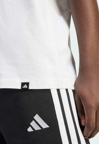 Biela bavlnená tričko s čiernym logom Adidas na lemovaní, spárovaná s čiernymi nohavicami so tromi bielymi pruhmi a logom.