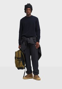 Marineblauer Pullover, dunkle Hose mit Bindeband in der Taille, braune Stiefel und ein olivgrüner Rucksack mit schwarzen Akzenten. Einfarbiger grauer Hintergrund.