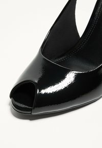 Fekete peep-toe magassarkú cipő, fényes lakkozott bőrből készült, ívelt dizájnnal és sima textúrával. A hangsúly a elülső nyitáskán.