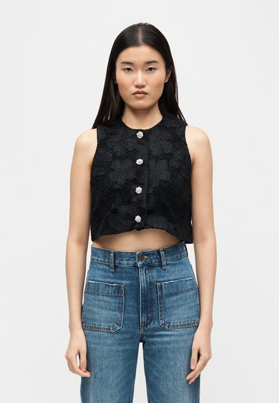 Γυναίκα που φοράει αμάνικο μαύρο floral δαντελένιο crop top με διακοσμητικά κουμπιά και ψηλόμεσο μπλε τζιν με μεγάλες μπροστινές τσέπες.