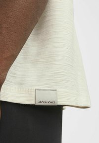 Svjetlosiva teksturirana majica s oznakom na rubu "JACK & JONES." Crna tkanina vidljiva ispod majice.