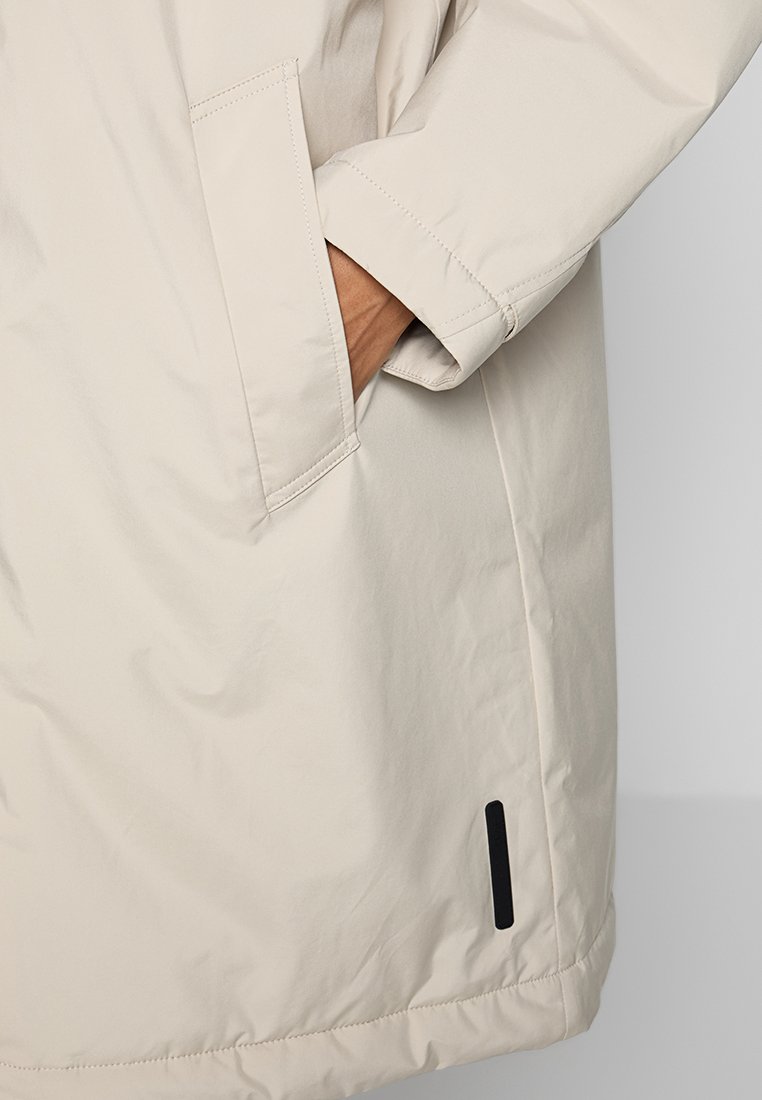 Main dans la poche d'un manteau beige à manches longues avec des coutures visibles et un petit détail rectangulaire noir près de l'ourlet inférieur.