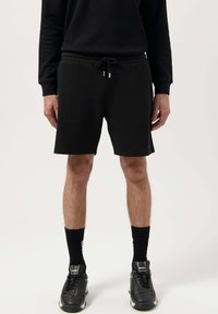 Black cotton shorts with an elastic waistband and drawstring, featuring a relaxed fit and above-knee length. Worn with black socks and sneakers.