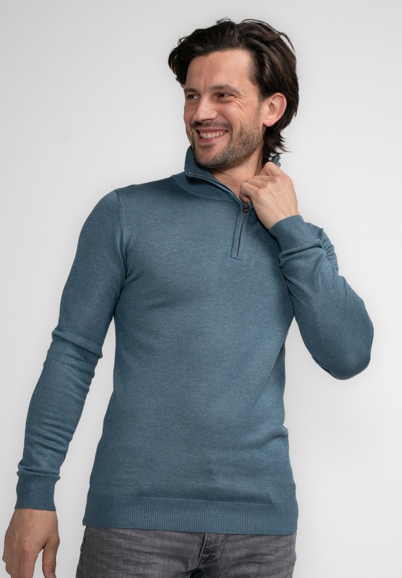 Blauwe gebreide pullover trui met een ritskraag, lange mouwen en geribde manchetten en onderrand. Het materiaal lijkt zacht en gestructureerd.