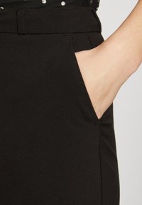 Pantalon noir sur mesure avec une poche latérale. Le tissu semble lisse avec une texture subtile. La taille est soigneusement finie sans embellissements.