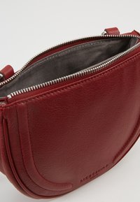 Czerwona skórzana torebka o zaokrąglonym kształcie, z zamkiem błyskawicznym u góry i szarą podszewką. Wykończenie zdobią charakterystyczne przeszycia.