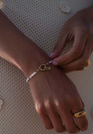 Eine Hand mit mittlerem Hautton, die einen goldenen Ring trägt, justiert ein zartes Perlenarmband mit goldenem Verschluss über einem strukturierten cremefarbenen Stoffhintergrund.