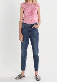 Canotta rosa senza maniche con colletto arricciato, abbinata a jeans blu a vita alta. Le calzature presentano dettagli metallici lucidi.