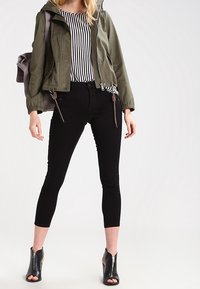 Olijfgroene cropped jas met trekkoord, zwarte nauwsluitende broek en zwarte open teen laarsjes. Witte en zwarte gestreepte blouse eronder.