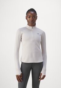 T-shirt à manches longues en beige clair, col zippé, tissu texturé avec des rayures verticales, logo sur la poitrine, associé à un legging noir.
