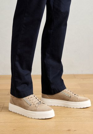 Beige Wildleder-Sneaker mit seitlichen Reißverschlüssen und weißen dicken Sohlen, getragen zu dunkelblauen, gerade geschnittenen Hosen auf Holzfußboden.