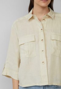 Kurzärmliges, geknöpftes Shirt aus hellem gelben Leinen mit zwei Brusttaschen, einem Kragen und umgekrempelten Ärmeln. Texturiert, leichtes Gewebe.
